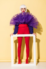 Full length view of woman in white wig holding frame on yellow background