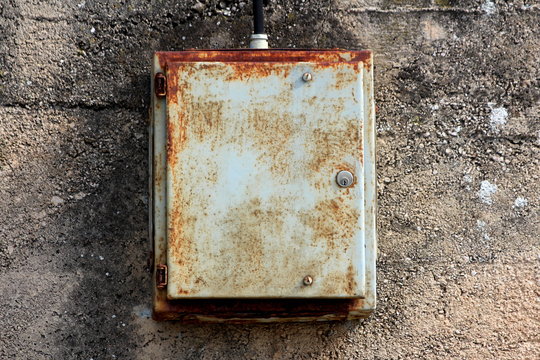 Partially rusted dilapidated locked metal electrical box mounted on cracked aged concrete wall connected with thick black electrical wire on warm spring day