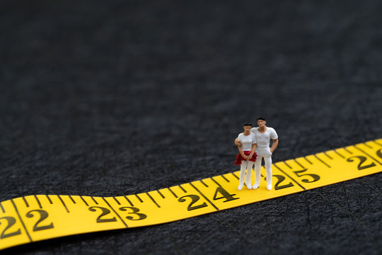 Young Adult, Gen Y Or New Age People Concept, Miniature People Couple In Casual White Cloth Standing On Number 25 On Yellow Measuring Tape Metaphor Of Their Age