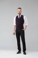 An elegant man in a vest and tie stands in full growth on a gray background.
