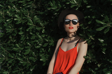 The girl in the summer in a black hat and sunglasses stands in the foliage of a green bush