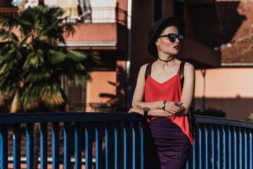 The girl in the summer in a black hat and sunglasses stands on the bridge against the backdrop of palm trees and houses in a European city. Travel and Vacation
