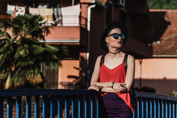 The girl in the summer in a black hat and sunglasses stands on the bridge against the backdrop of palm trees and houses in a European city. Travel and Vacation