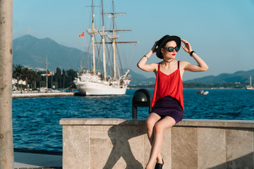 A girl in sunglasses and a black hat sits against the backdrop of the sea and the ancient ship in Europe. Travel and Vacation