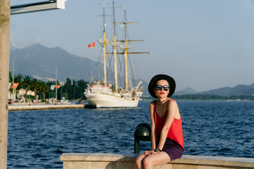A girl in sunglasses and a black hat sits against the backdrop of the sea and the ancient ship in Europe. Travel and Vacation