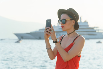 Girl in sunglasses and a black hat on the background of yachts in the resort European city takes pictures on a smartphone or takes a selfie. Travel and Vacation