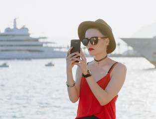 Girl in sunglasses and a black hat on the background of yachts in the resort European city takes pictures on a smartphone or takes a selfie. Travel and Vacation