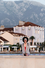 A girl in sunglasses and a black hat is standing on a pier near the sea in a European resort town on the background of luxury houses and palm trees. Travel and Vacation
