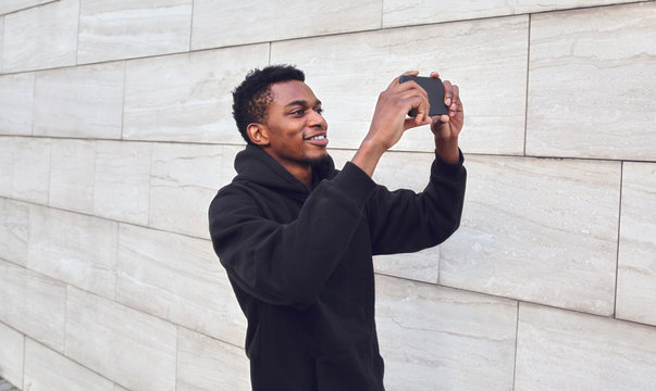 Cheerful smiling african man taking selfie picture by phone on city street over gray wall background
