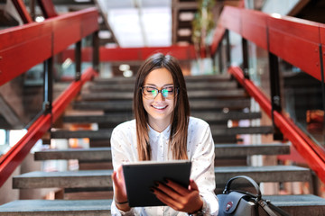 Close up of smiling female student with eyeglasses and brown hair using tablet while sitting on the stairs. Next to her bag.