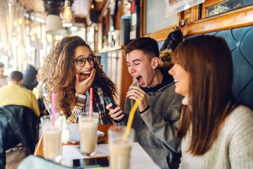 Three laughing multicultural classmates sitting at coffee shop, drinking coffee and looking at smart phone. On desk notebooks, markers and pens.