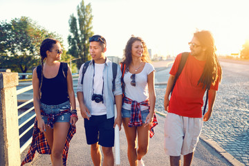 Obraz premium Four cheerful multicultural tourists with backpacks on backs walking down the street and chatting. Summertime vacations concept.