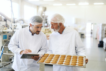 Worker holding casserole with cookies while supervisor checking quality and holding tablet. Food factory interior.