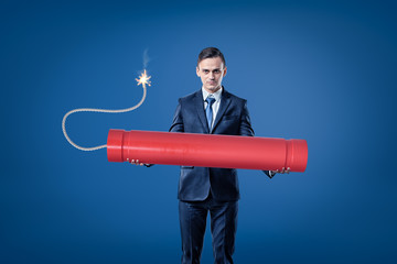 Young businessman holding big red tnt dynamite stick with lighted fuse on blue background
