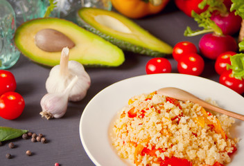 Delicious homemade vegetarian couscous with tomatoes, carrots, pepper and fresh basil on a dark kitchen table with vegetables on the background