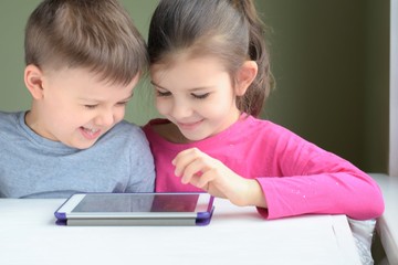 White caucasian toddler boy and beautiful girl playing together on tablet games. Portrait of smiling little brother and sister. Children play a computer game on the tablet