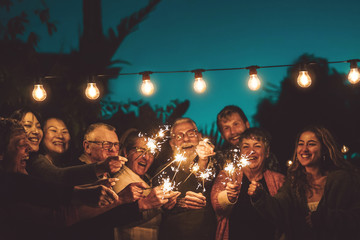 Happy family celebrating with sparkler at night party outdoor - Group of people with different ages and ethnicity having fun together outside - Friendship, eve and celebration concept - obrazy, fototapety, plakaty