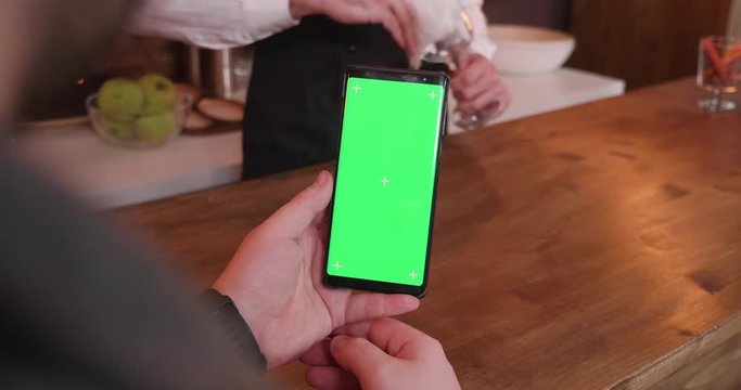 Man swipes on a phone with green screen while sitting at the barcounter and the bartender is cleaning a glass in the background