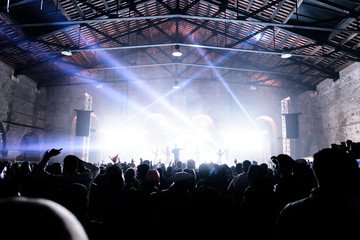 People on the dance floor during a concert in a hangar. A great glow covers the singer and the artistic animation on the stage. Laser and lights illuminate the party. Wide view. Hands up in the air