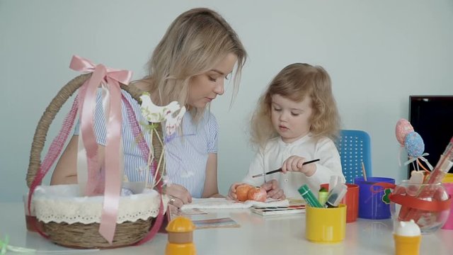 Happy Easter! Mother and her little daughter painting Easter eggs at the festive table with basket and eggs. Slow Motion