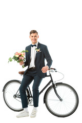 cheerful groom standing near bike and holding wedding bouquet while looking at camera isolated on white