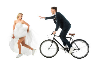 pretty bride in wedding dress and sneakers running from groom on bicycle isolated on white