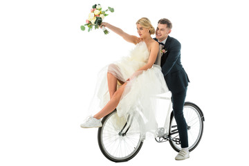 beautiful bride holding wedding bouquet in raised hand while sitting on bicycle together with groom...
