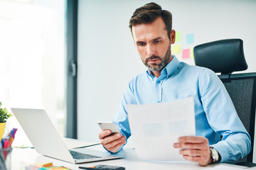 A man sitting at the desk at the office, checking financial documents and holding smartphone - obrazy, fototapety, plakaty