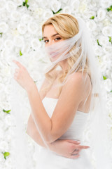 attractive young bride hiding face behind bridal veil while posing at camera on white floral background