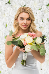 Naklejka premium beautiful young woman holding wedding bouquet and looking at camera on white floral background