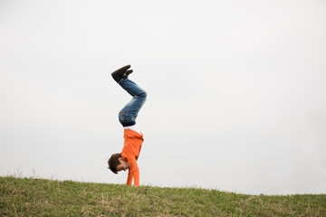 active little caucasian boy in jeans standing on hands upside down on green grass outdoors with copy space