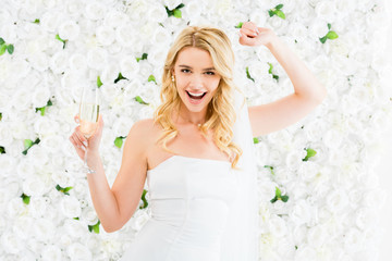 Obraz na płótnie Canvas cheerful young bride with glass of champagne looking at camera on white floral background