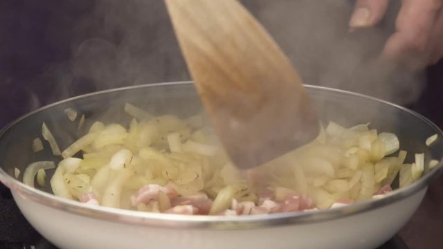  Stirring onion with bacon in hot pan, hands cooking it with a wooden spatula