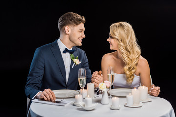 happy young groom and bride holding hands while sitting at served table isolated on black