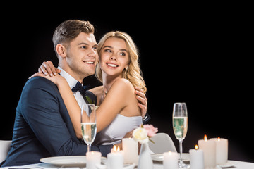 happy groom and bride embracing while sitting at served table isolated on black