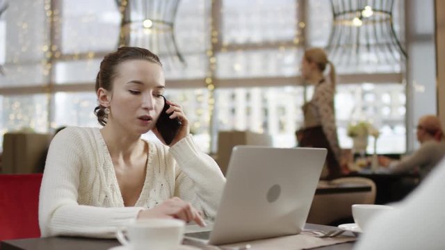 Young woman is talking over the smartphone while sitting by table in front of laptop and looking to man sitting oposite of her