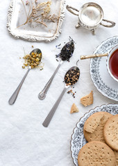Different types of tea on vintage metal spoons on white fabric background, teacup, dried flower, silver plate, cookies. Vintage food and drink setting styling.  Organic healthy well-being lifestyle.