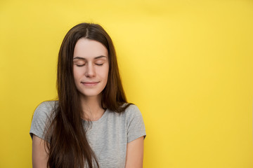 Fototapeta premium Close up shot of pleased adorable young female with wears gray T-shirt, has soap smile, keeps eyes closed, dreams about something pleasant. Attractive woman poses indoor on yellow will