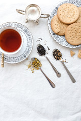 Different types of tea on vintage metal spoons on white fabric background, teacup, dried flower, silver plate, cookies. Vintage food and drink setting styling.  Organic healthy well-being lifestyle.