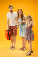 Fototapeta premium Sad unhappy parent with daughter and suitcase at studio isolated on yellow background. Travel, vacation, parenthood, togetherness, tourism concept.