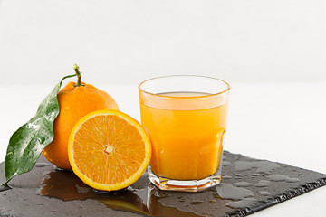 An inviting glass full of orange juice, an half orange and a whole one with leaf on a wet square slate plate onwhite background