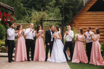 Groom and bride stand with groomsman and bridesmaid outside. Newlyweds kissing and friend clap....