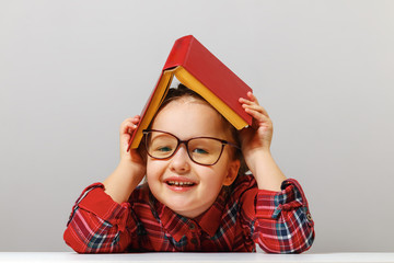 Funny girl with a book on her head and wearing glasses is sitting at the table, looking at the...