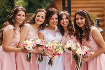 Happy bride with bridesmaid hold bouquets and have fun outside. Beautiful bridesmaid in same dresses stand by the charming bride in long wedding dress