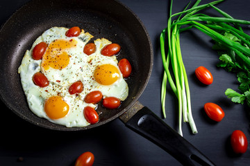 Fried eggs with tomatoes in a pan and fresh onions on dark background . Top view.