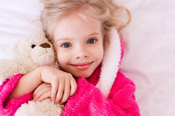 Smiling kid girl 4-5 year old holding teddy bear lying in bed. Looking at camera. Childhood.