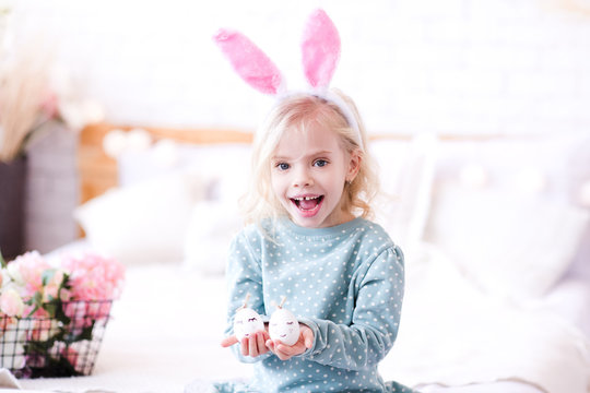Happy kid girl 4-5 year old holding Easter eggs with cute faces in room. Wearing bunny headband. Looking at camera.