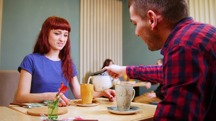 A couple sitting by table in a cafe. A man pours the drink in the cups