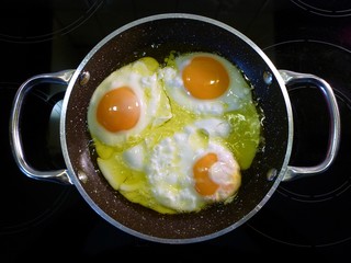 Closeup top view of cooked homemade meal or breakfast of three fried eggs in a frying pan in hot extra virgin Greek olive oil ready to serve isolated on a black ceramic glass range cooktop