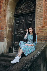 Attractive young smiling woman sitting near old building in cute summer dress with flower print